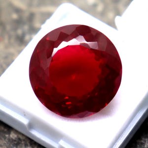 Puede incluir: Una gema facetada, redonda, de color rojo intenso, posiblemente un rubí, descansa en una caja de presentación de plástico blanco. La gema es el punto focal, mostrando su color y corte brillantes. El fondo es una superficie de piedra rugosa y gris.