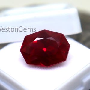 Puede incluir: Una gema facetada de color rojo intenso, posiblemente un rubí, descansa en una caja de plástico blanca. La gema tiene forma octogonal y refleja la luz, mostrando su talla brillante. El texto "WestonGems" es visible.
