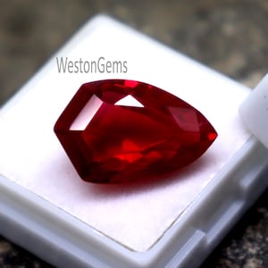 May include: A faceted, deep red gemstone presented in a white plastic case. The gem has a brilliant cut, reflecting light. The text "WestonGems" is visible on the case. The gemstone is a vibrant, eye-catching piece.