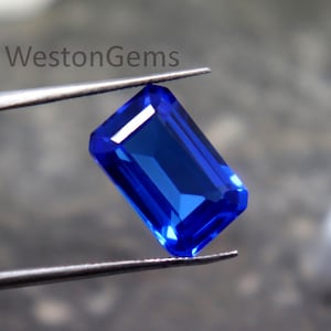 Puede incluir: Una gema azul zafiro, rectangular y brillante, es sostenida por unas pinzas. La gema tiene un corte facetado, que refleja la luz. La imagen está sobre un fondo borroso y neutro. El texto "WestonGems" es visible en la esquina superior izquierda.