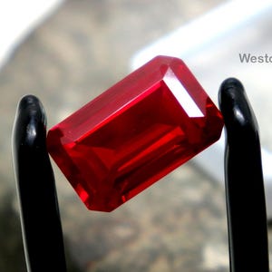 Puede incluir: Una gema roja brillante, rectangular, es sostenida por unas pinzas negras. La gema está facetada, reflejando la luz. Un recipiente cuadrado blanco y una superficie de piedra texturizada están en el fondo. El texto "WestonGems" es visible.