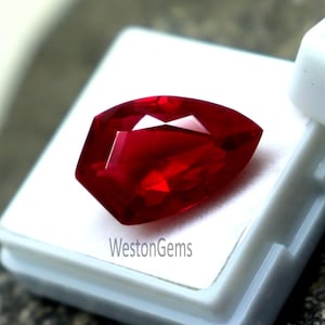 May include: A faceted, deep red gemstone with a pear-like shape rests in a white plastic case. The gem is highly reflective, showcasing multiple facets. The text "WestonGems" is printed on the case. The background is a textured, dark surface.