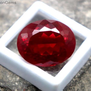 Può includere: Una gemma rosso intenso, sfaccettata e di forma ovale, presentata in una cornice quadrata bianca. La gemma è posta su una superficie di pietra grigia e ruvida. Il testo "WestonGems" è visibile.