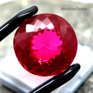 Puede incluir: Una gema facetada, redonda, de color rojo intenso, sujeta con pinzas negras. La gema está colocada sobre una base blanca sobre un fondo gris texturizado. El texto "WestonGems" es visible en la esquina superior derecha.