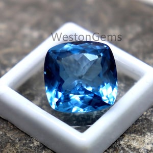 Puede incluir: Una gema azul brillante, tallada en cojín, colocada en un estuche cuadrado blanco. La gema tiene múltiples facetas que reflejan la luz, creando un efecto brillante. El texto "WestonGems" es visible.