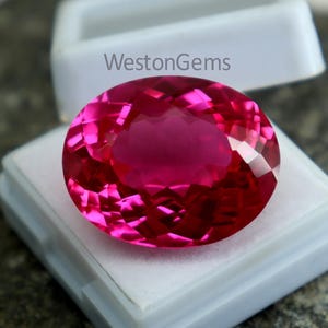 May include: A large, faceted, oval-shaped, hot pink gemstone is displayed in a white plastic case. The gemstone has a brilliant cut, reflecting light. The text "WestonGems" is visible on the case.