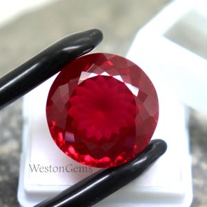 Puede incluir: Una gema facetada, redonda, de color rojo intenso, sostenida por unas pinzas negras. La gema está colocada sobre una superficie blanca dentro de un recipiente blanco. El texto "WestonGems" es visible.