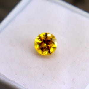 May include: A round, faceted yellow gemstone with a brilliant cut, displayed on a white square display pad inside a small white plastic container. The stone exhibits a vibrant, translucent golden hue with visible internal light reflections.