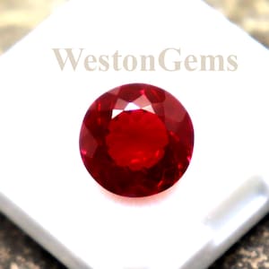 May include: A vibrant, round, deep red gemstone, possibly a ruby, sits on a white display case. The case is open, showcasing the gem's cut. The text "WestonGems" is printed on the case. The background is a textured, dark surface.