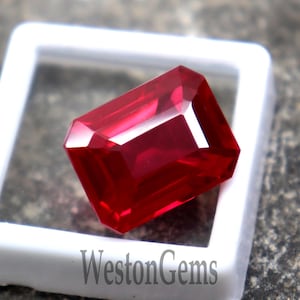 Puede incluir: Un rubí facetado, rectangular, en un expositor blanco. El rubí es de color rojo intenso y tiene un brillo brillante. El expositor está sobre una superficie de piedra gris texturizada. El texto "WestonGems" es visible.