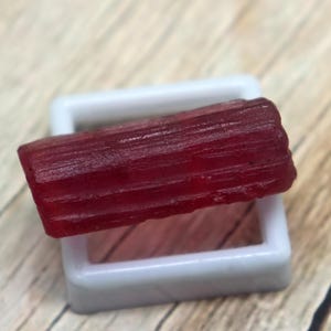 May include: A raw, deep red gemstone, possibly a tourmaline, rests in a white plastic display case. The gemstone has a rectangular shape with visible striations and a slightly translucent appearance. The background is a wooden surface.