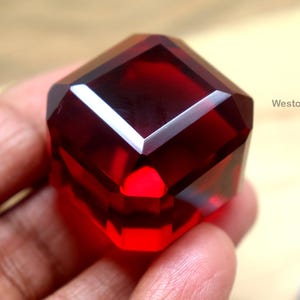 Puede incluir: Una gema facetada de color rojo intenso con forma geométrica. La gema se sostiene en una mano, mostrando sus superficies pulidas y reflejos internos. El texto "WestonGems" es visible en el fondo.