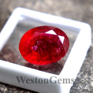 May include: A faceted, oval-shaped, red gemstone is displayed in a white plastic case. The gemstone has a brilliant, reflective surface. The case is set on a textured, gray stone surface. The text "Weston Gems" is visible on the case.