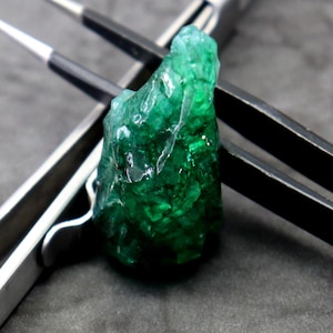 Può includere: Uno smeraldo grezzo di colore verde intenso posizionato tra due pinzette di metallo. Una pinzetta è color argento, mentre l'altra è nera con punte argentate. Lo smeraldo presenta una consistenza cristallina irregolare con sfaccettature traslucide.