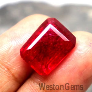 May include: A vibrant, rectangular-cut red gemstone with faceted surfaces that reflect light. The stone displays a deep, saturated crimson hue and is positioned against a blurred background. The text WestonGems is visible on the surface.