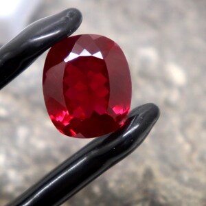 Puede incluir: Una piedra preciosa de color rojo intenso con talla cojín, sostenida por unas pinzas negras. La piedra presenta una superficie facetada que refleja la luz, mostrando un color carmesí vibrante y saturado en todo su cuerpo transparente.