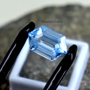 Puede incluir: Una piedra preciosa de color azul claro con corte esmeralda y superficie facetada, sostenida por unas pinzas negras. La piedra es transparente, refleja la luz y muestra una forma rectangular clara. El texto WestonGems es visible en negro.