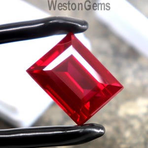 May include: A vibrant red gemstone with a rectangular emerald cut, held by black metal tweezers. The stone features sharp, clean facets that reflect light, creating a deep crimson hue. The text WestonGems is visible in the upper center area.