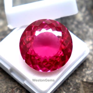 May include: A faceted, round, bright pink gemstone displayed in a white plastic case. The gem has a high degree of clarity and brilliance, reflecting light. The case is open, revealing the gem's polished surface. The gemstone is a vibrant shade of pink.
