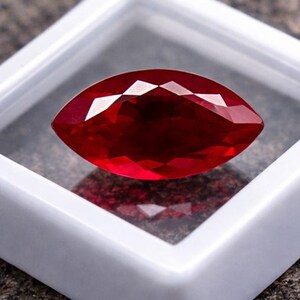 Puede incluir: Una gema roja intensa, tallada en forma de marquesa y facetada, se muestra en una caja de plástico blanco. La gema está sobre una superficie texturizada de color gris oscuro. El texto "WestonGems" es visible.