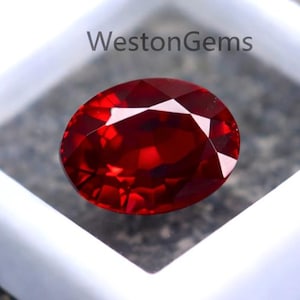 Può includere: Una vivace gemma rossa, taglio ovale, esposta in un astuccio quadrato bianco. La gemma ha una superficie sfaccettata brillante, che riflette la luce. L'astuccio riporta la scritta "WestonGems". La gemma è posta su una superficie grigia strutturata.