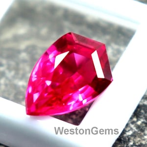 Könnte beinhalten: Ein facettierter, leuchtend roter Edelstein wird in einem weißen, quadratischen Behälter präsentiert. Der Edelstein hat einen einzigartigen Schliff mit mehreren Facetten. Der Behälter liegt auf einer strukturierten, grauen Oberfläche. Der Text "WestonGems" ist sichtbar.