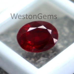 Peut inclure: Une pierre précieuse rouge foncé, taillée en ovale, présentée dans un contenant carré blanc. La gemme est taillée à facettes, reflétant la lumière. Le contenant est posé sur une surface grise texturée. Le texte "WestonGems" est visible.