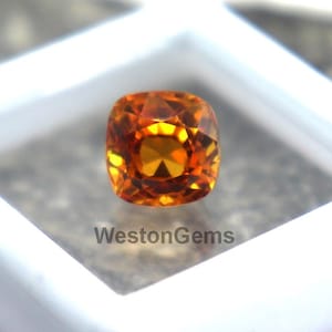 Puede incluir: Una gema naranja, tallada en cojín, se exhibe dentro de una vitrina blanca cuadrada. La gema tiene múltiples facetas que reflejan la luz. La vitrina está sobre una superficie gris texturizada. El texto "WestonGems" es visible.