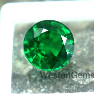 May include: A round, faceted, vibrant green gemstone, set in a white square display case. The gemstone is a brilliant emerald color, with visible facets catching the light. The text "WestonGems" is visible on the case.