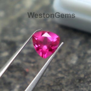 Puede incluir: Una gema rosa vibrante en forma de corazón es sostenida por unas pinzas sobre una superficie gris texturizada. La gema tiene un corte brillante, reflejando la luz. Un contenedor blanco está en el fondo. El texto "WestonGems" es visible.