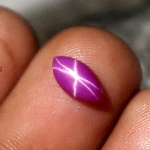 Puede incluir: Una gema ovalada de color rosa púrpura con un patrón de estrella blanca. La gema pulida se sostiene entre los dedos, mostrando su efecto de estrella. La gema es un rubí estrella, un hermoso ejemplo de piedra preciosa.