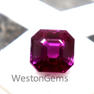 Puede incluir: Una gema facetada de color magenta con corte cuadrado, colocada sobre una superficie blanca. La gema se muestra en una caja blanca, con el texto "WestonGems" visible. Las facetas de la gema reflejan la luz, mostrando su brillo.