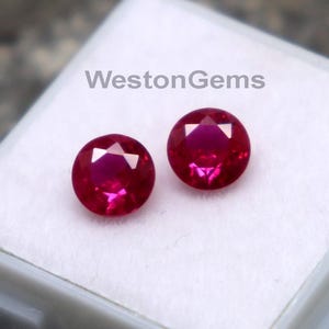 May include: Two round, faceted, deep red gemstones are displayed in a white plastic case on a textured, dark gray surface. The gemstones are a vibrant shade of red, and the case has the text "WestonGems" printed on it.