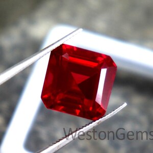 Könnte beinhalten: Ein quadratisch geschliffener, roter Edelstein wird von einer Pinzette gehalten. Der Edelstein hat eine tiefe, rote Farbe und scheint facettiert zu sein. Der Text "WestonGems" ist auf dem weißen Hintergrund sichtbar.