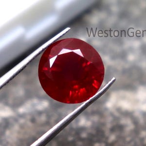 Puede incluir: Una gema redonda y facetada de color rojo intenso es sostenida por unas pinzas plateadas. Se muestra sobre una superficie gris texturizada. Una caja blanca se encuentra en el fondo. La gema es un rubí pulido.