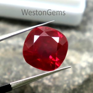 Puede incluir: Una vibrante gema roja con talla cojín es sostenida por unas pinzas contra un fondo texturizado. La gema tiene una superficie facetada que refleja la luz, mostrando su brillo. El contenedor blanco en el fondo tiene el texto "WestonGems".