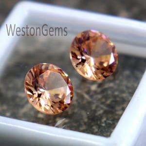 Puede incluir: Dos gemas ovaladas de color melocotón exhibidas en un contenedor cuadrado blanco. Las gemas tienen un corte brillante, reflejando la luz. El contenedor está sobre un fondo oscuro y texturizado. El texto "WestonGems" es visible.