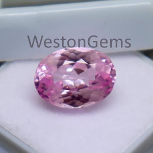 Puede incluir: Una gema rosa de corte ovalado, exhibida en una caja de plástico blanco. La gema tiene múltiples facetas, reflejando la luz. La caja está abierta, revelando la gema sobre un fondo blanco. El texto "WestonGems" es visible.