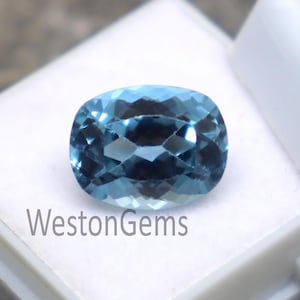 Puede incluir: Una gema azul claro, tallada en forma ovalada, se encuentra en una caja blanca. La gema tiene múltiples facetas que reflejan la luz. El texto "WestonGems" está impreso en la caja. La piedra preciosa es de un tono azul vibrante.