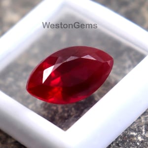 May include: A marquise-cut, deep red gemstone is displayed in a white plastic case. The gemstone is faceted, reflecting light. The case sits on a rough, gray stone surface. The text "WestonGems" is visible on the case.