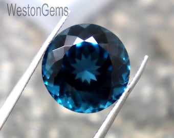 Puede incluir: Una gema redonda facetada de color verde azulado oscuro, sujeta con pinzas plateadas. La gema tiene un patrón de estrella en el centro. El texto "WestonGems" es visible en el fondo. La gema es de talla brillante.