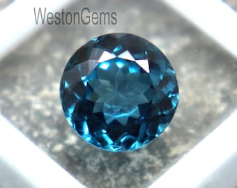 Puede incluir: Una gema redonda, facetada, de color verde azulado se muestra dentro de un contenedor cuadrado de plástico blanco. La gema tiene un corte brillante, reflejando la luz. El contenedor está sobre una superficie de piedra gris texturizada. El texto "Westongems" es visible.