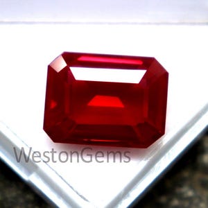 May include: A vibrant, rectangular-cut, deep red gemstone, possibly a ruby, is displayed in a white plastic case. The gem has a polished surface and is set against a neutral background. The text "WestonGems" is visible on the case.