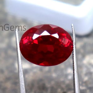 Puede incluir: Una gema roja intensa, facetada y de forma ovalada, es sostenida por unas pinzas plateadas. La gema tiene múltiples facetas reflectantes. En el fondo, una caja blanca. El texto "WestonGems" se aprecia en la esquina superior izquierda.