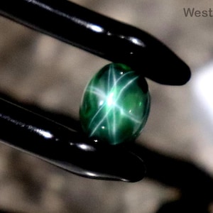 Pode incluir: Uma gema verde oval polida com um padrão de estrela de seis pontas, segurada por pinças pretas. O fundo é neutro. O texto "WestonGems" é visível no fundo.