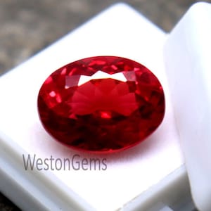 May include: A faceted, oval-shaped, vibrant red gemstone sits in a white plastic display case. The gem is set against a textured, grey stone surface. The text "WestonGems" is visible on the case.