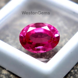 Op de afbeelding: Een levendige, ovaal geslepen, roze edelsteen gepresenteerd in een witte, vierkante plastic behuizing. De edelsteen is geplaatst op een getextureerd, grijs stenen oppervlak. De tekst "WestonGems" is zichtbaar.