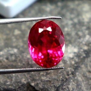 May include: A faceted, oval-shaped, vibrant red gemstone is held by tweezers. The gem has a brilliant cut, reflecting light. The background is a rough, gray stone surface. The text "WestonGems" is visible in the upper left corner.