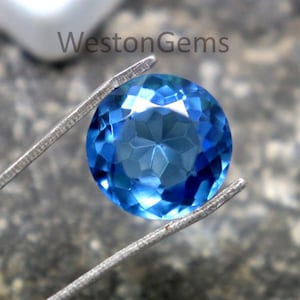 Puede incluir: Una gema azul redonda y facetada, sujeta por unas pinzas plateadas. La gema es de un tono azul vibrante, con múltiples facetas que captan la luz. El fondo es una superficie de piedra gris texturizada. El texto "WestonGems" es visible.