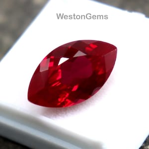 May include: A marquise-cut, vibrant red gemstone rests on a white display case. The gemstone is faceted, reflecting light and showcasing its deep color. The display case is set against a textured, gray stone surface. The text "WestonGems" is visible on the case.